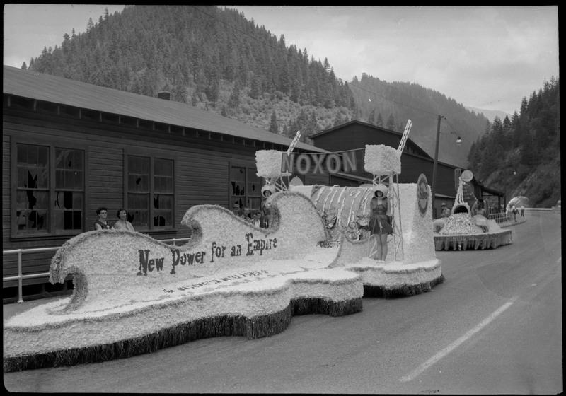Two women standing on the Noxon float during the Silver Jubilee. Bystanders watch from the sidewalk. Text along the side of the float reads, "New Power for an Empire."