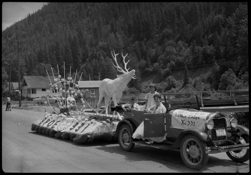 A woman and two children sitting in a vehicle with a poster that reads, "Old Timers Wallace Lodge No. 331." The float behinds the vehicle is decorated with a fake moose and flowers. The side of the float reads, "BPOE 331."