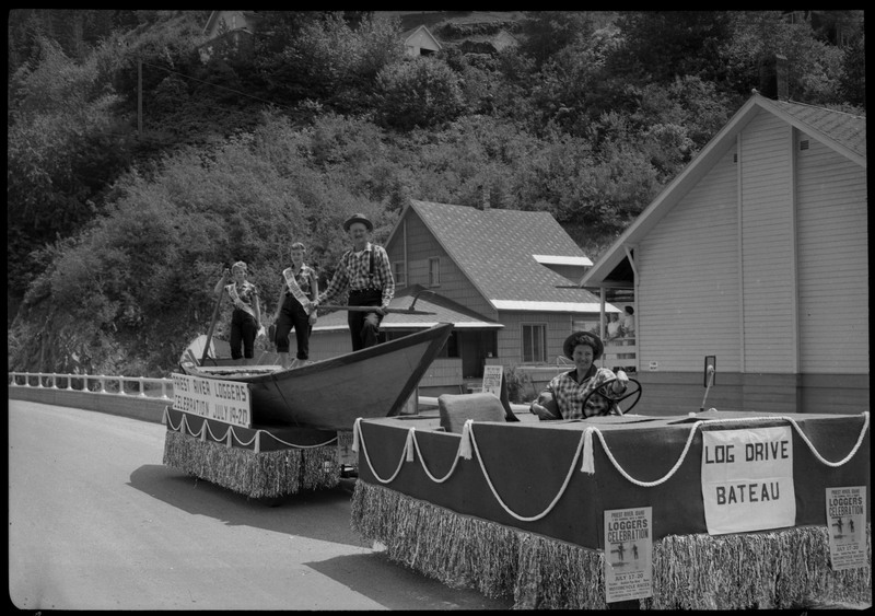 Four people on a float shaped like a boat during the Silver Jubilee. Signs on the float read, "Priest River Loggers Celebration July 19-20" and "Log Drive Bateau." There are also smaller posters advertising the Loggers Celebration.