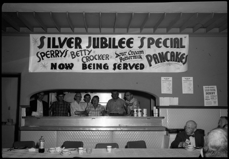 Seven people behind a counter and beneath a poster reads, "Silver Jubilee "Special" Pancakes Sperry's Betty Crocker Sour Cream Buttermilk Now Being Served." Some people can be seated on the right corner.