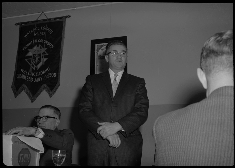 A man wearing a suit stands during a Knights of Columbus Jubilee event. Another man sits on his left and the back of a man can be seen on the right. A banner for the Wallace Knights of Columbus is hanging on the wall behind all three men.