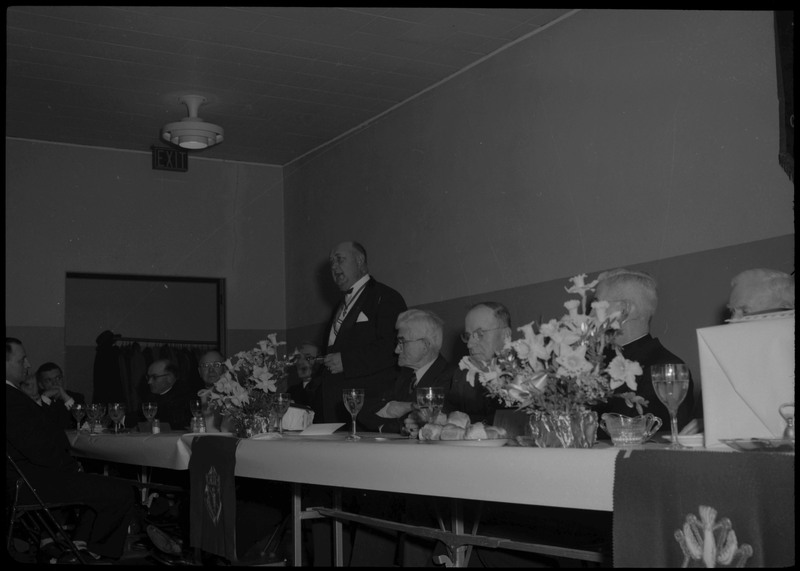A man stands and speaks at a table with other seated people during a Knights of Columbus Jubilee event.