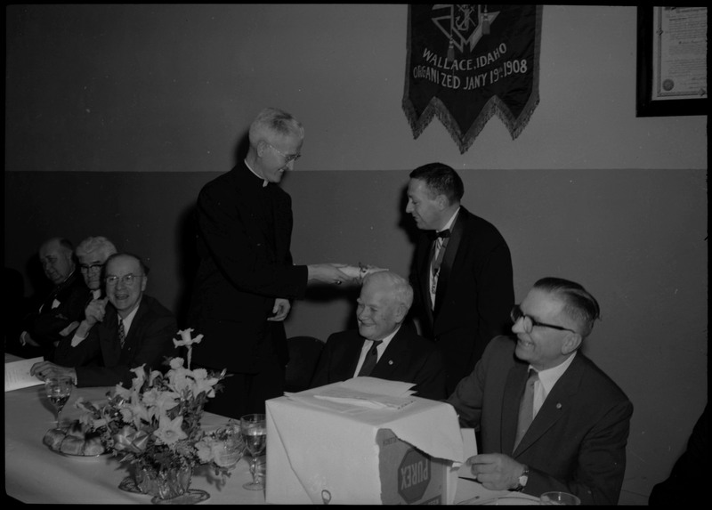 Two men shaking hands and exchanging a roll of paper during a Knights of Columbus Jubilee event. Other people can be seen around them.