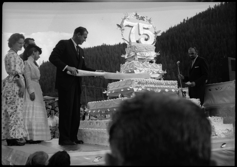 A man holding a large knife to cut a huge cake during the Silver Jubilee. The cake is decorated with the number "75".
