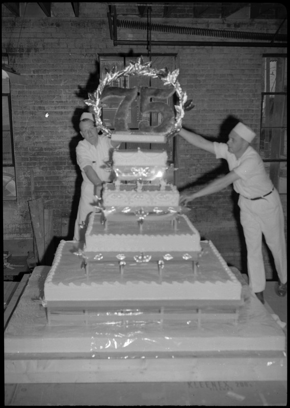 Two men wearing matching uniforms and hats work on making a huge cake during the Silver Jubilee. The cake is decorated with the number, "75".