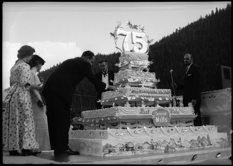 A man cutting a huge cake during the Silver Jubilee with a smaller knife. The cake is decorated with the number "75" and the text, "Happy Birthday Wallace, Silver Jubilee" "Best Wishes from General Mills." Other people are also standing and watching near the cake.
