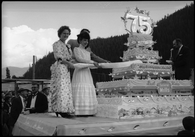 Three people holding a large knife to cut a huge cake during the Silver Jubilee. The cake is decorated with the number "75" and the text, "Happy Birthday Wallace, Silver Jubilee" "Best Wishes from General Mills."