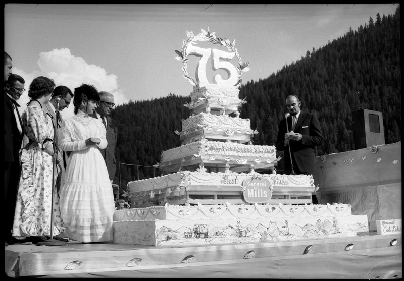 People standing on a stage next to a large cake during the Silver Jubilee. A man speaks into a microphone on the right. The cake is decorated with the number "75" and the text, "Happy Birthday Wallace, Silver Jubilee" "Best Wishes from General Mills."