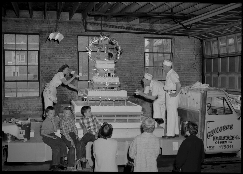 Two people use frosting bags to decorate the Silver Jubilee cake. The cake is on the flatbed of a Builders Hardware Co. truck. Three children and one adult are on the flatbed as three other people watch on the ground.