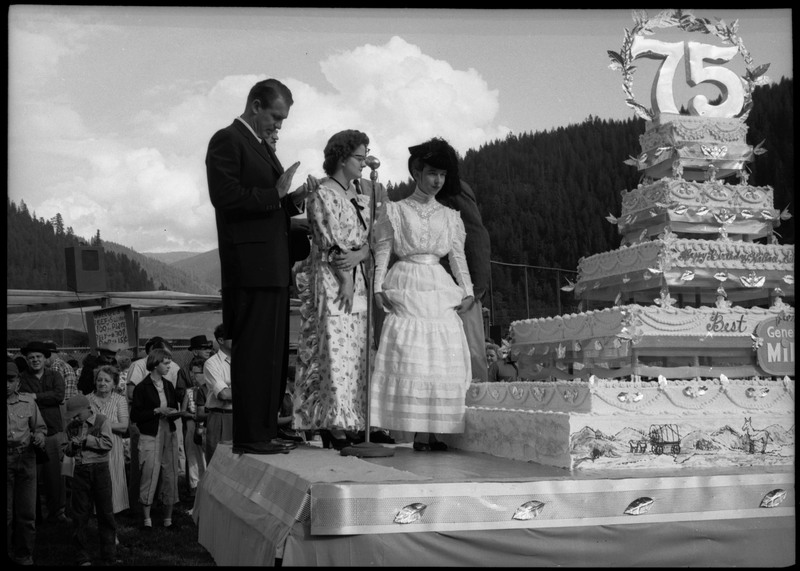 People standing on a stage next to a large cake during the Silver Jubilee. The cake is decorated with the number "75".