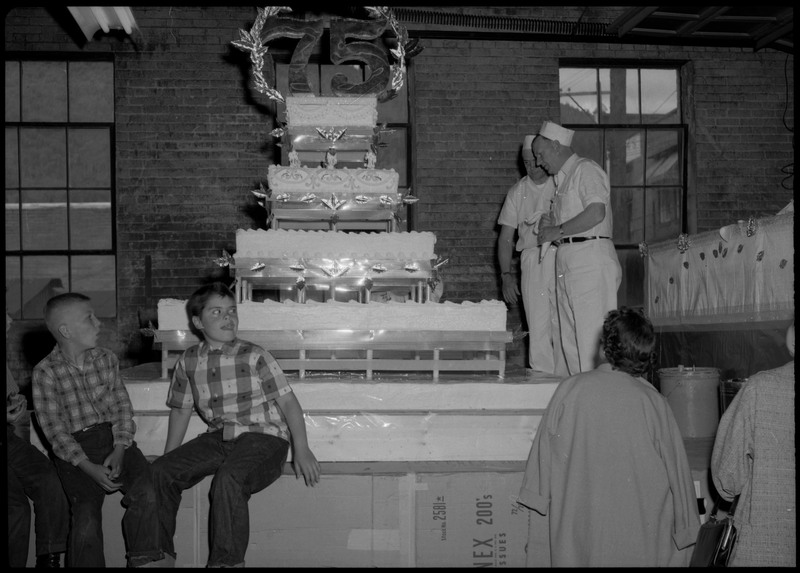 Two men, both wearing identical white uniforms and hats, stand near the Silver Jubilee cake. Three children are seated near the cake and two people stand watching the cake.