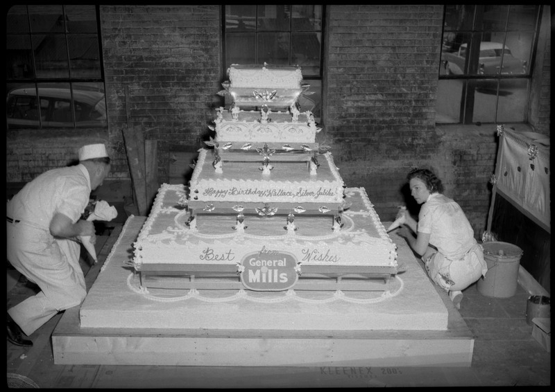 Two people use frosting bags to decorate the Silver Jubilee cake. The cake reads, "Happy Birthday, Wallace, Silver Jubilee" and "Best Wishes from General Mills."