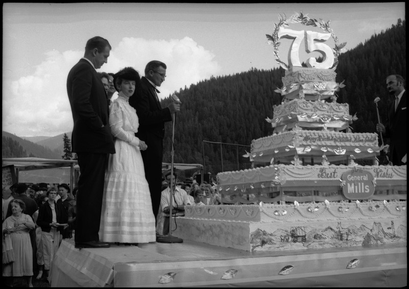 People standing on a stage with a large Silver Jubilee cake. Two of the people are holding microphones. Other people can be seen watching from the ground.
