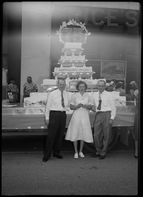 Two men and one woman pose in front of the Silver Jubilee cake. People can be seen behind the cake.