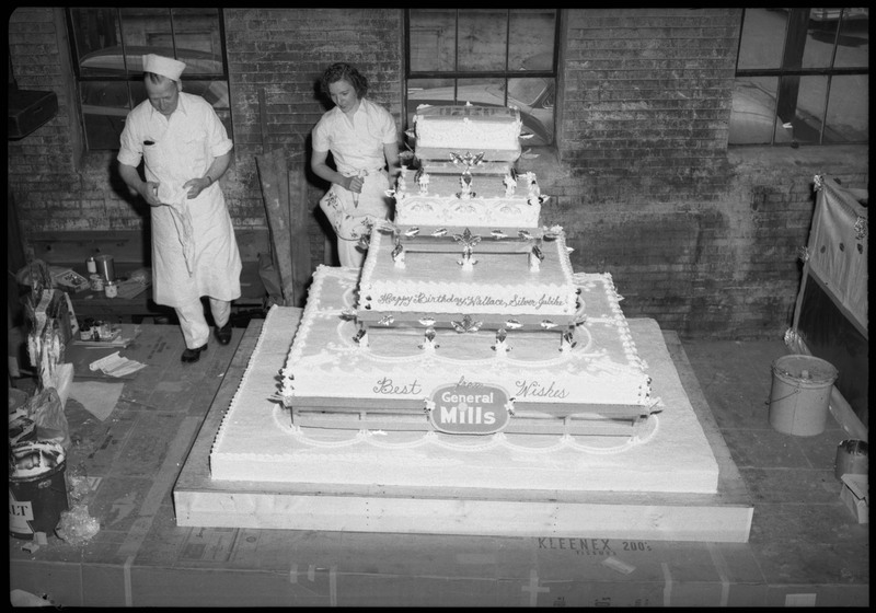 Two people holding frosting bags near the Silver Jubilee cake.