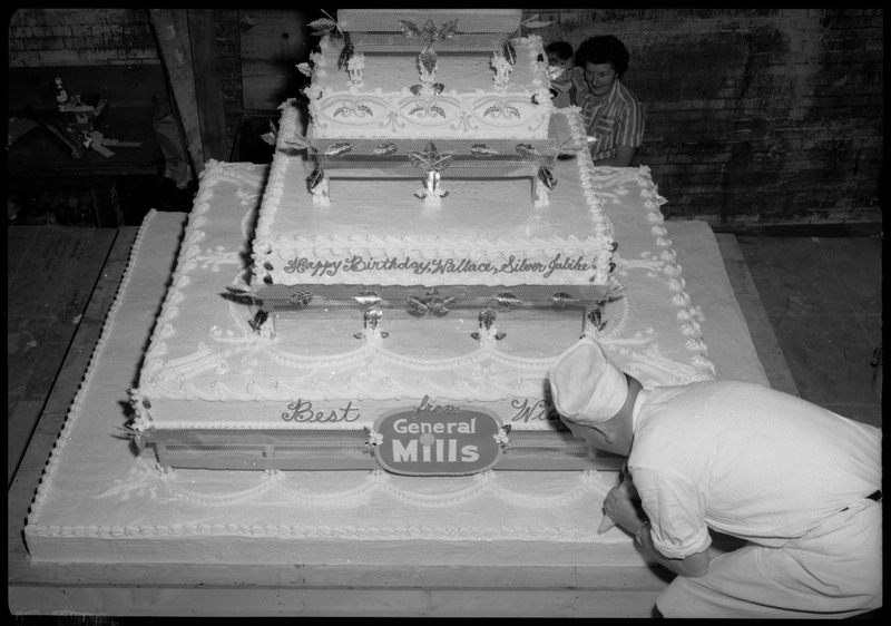 A man uses a frosting bag to decorate the Silver Jubilee cake. The cake reads, "Happy Birthday, Wallace, Silver Jubilee" and "Best Wishes from General Mills."