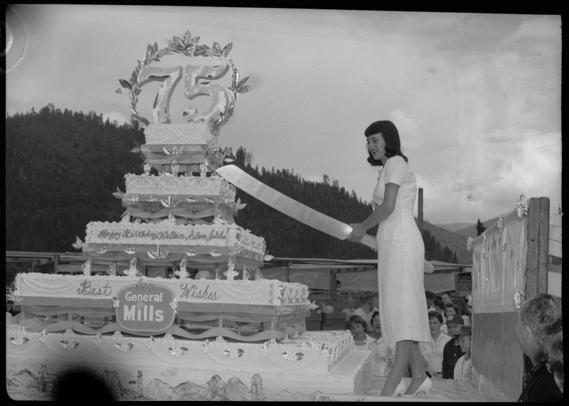 A woman holds a large knife to cut the Silver Jubilee cake. People are watching from the ground.