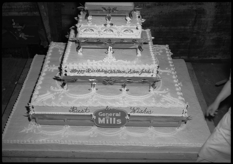 View of the Silver Jubilee cake. The cake reads, "Happy Birthday, Wallace, Silver Jubilee" and "Best Wishes from General Mills." Someone's arm and leg can be seen on the right.