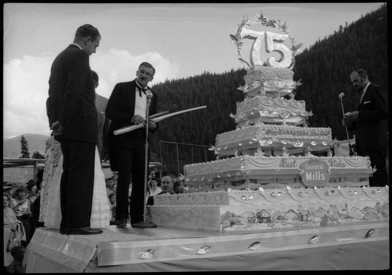 People standing on a stage next to the Silver Jubilee cake. One of the people is holding a large knife to cut the cake with. Other people are watching on the ground.