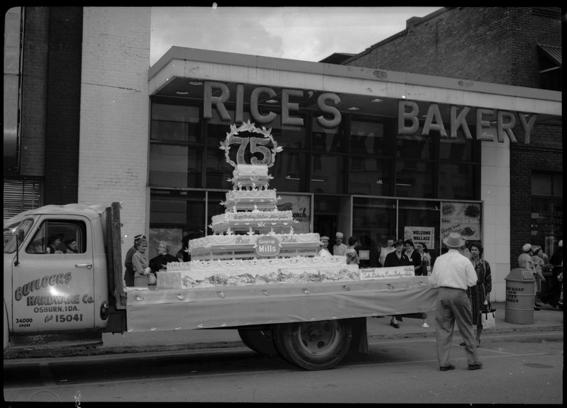The Silver Jubilee cake sitting on the flatbed of a truck. The truck is parked in front of Rice's Bakery. People can be seen standing on the sidewalk.