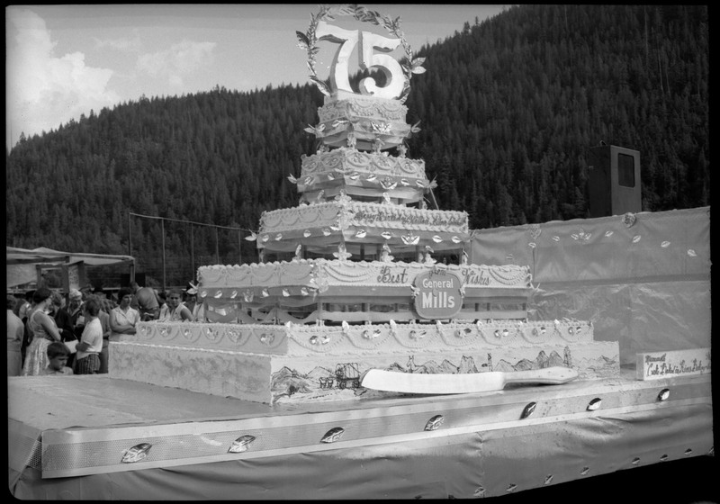 View of the Silver Jubilee cake. There is a large knife sitting in front of the cake The cake reads, "Happy Birthday, Wallace, Silver Jubilee" and "Best Wishes from General Mills." People can be seen in the background behind the cake.