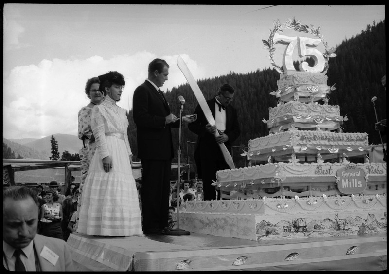People standing on a stage next to the Silver Jubilee cake. One of the people is holding a large knife to cut the cake with. Other people are watching on the ground.