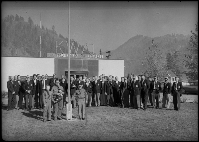 Men wearing suits gathered in front the KWAL The Voice of the Coeur D'Alenes building. Three boys are standing near a flagpole and one is holding a folded American flag.
