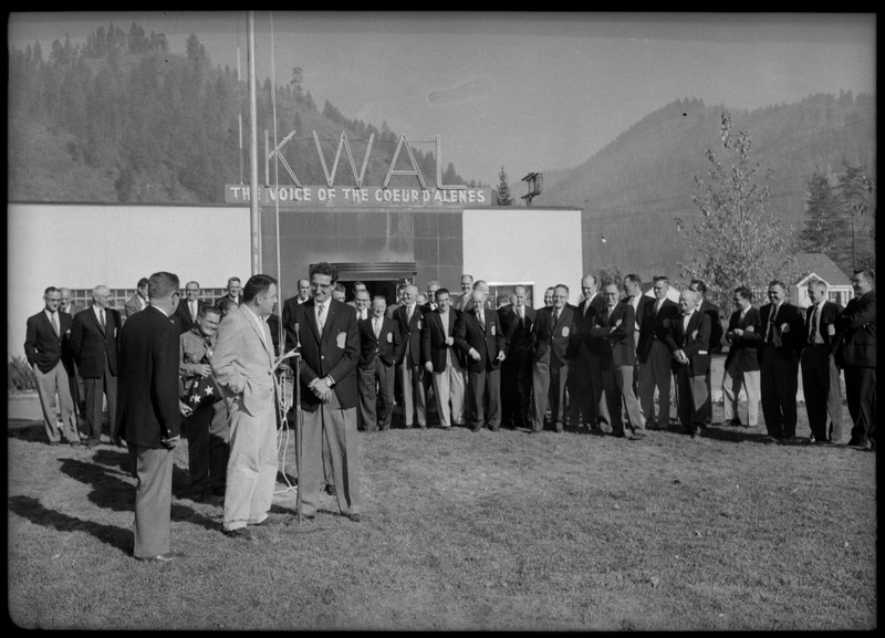Men wearing suits gathered in front the KWAL The Voice of the Coeur D'Alenes building. Three men are standing in front of the flagpole, one is speaking into a microphone.