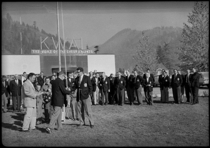 Men wearing suits gathered in front the KWAL The Voice of the Coeur D'Alenes building. Three men are standing near a microphone while young boys stand behind the three men near a flagpole.