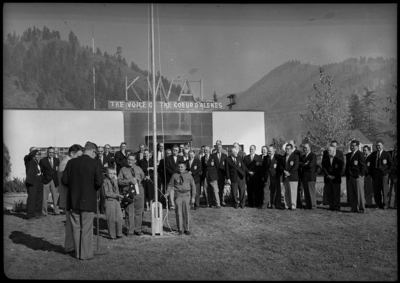 Men wearing suits gathered in front the KWAL The Voice of the Coeur D'Alenes building. Two men are standing near a microphone while three young boys stand besides a flagpole. One of the boys is holding a folded American flag.