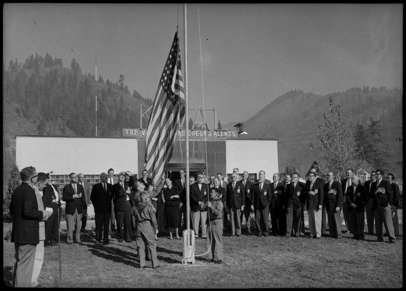 Men wearing suits gathered in front the KWAL The Voice of the Coeur D'Alenes building. Three boys are raising the American flag.