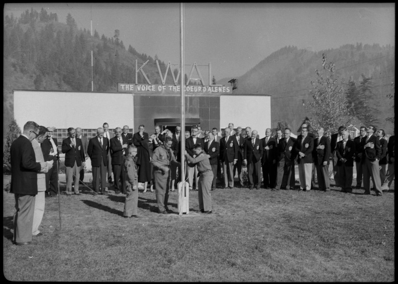 Men wearing suits gathered in front the KWAL The Voice of the Coeur D'Alenes building. Three boys are raising the American flag.