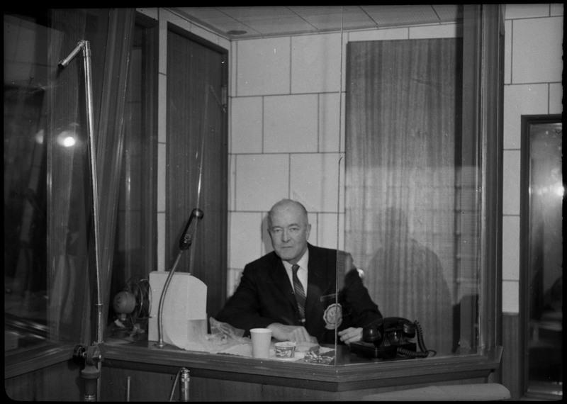 A man sitting in a booth at the KWAL broadcasting radio station. There is a microphone and a phone in the booth.