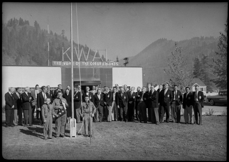 Men wearing suits gathered in front the KWAL The Voice of the Coeur D'Alenes building. Three boys are standing near a flagpole and one is holding a folded American flag.