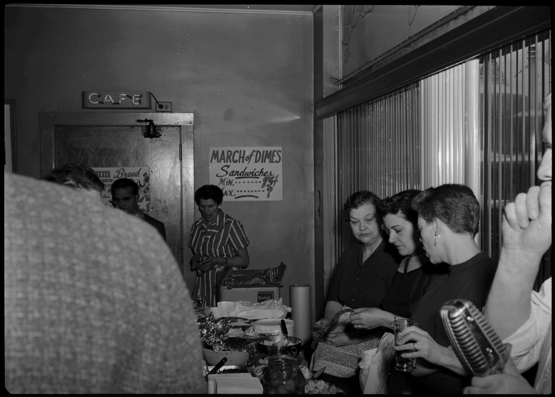 People near a table with food during the Albie's March of Dimes event. A sign on the back wall advertises the March of Dimes sandwiches.