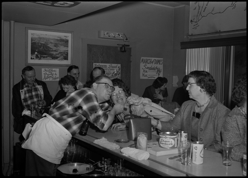 People sitting at a bar area during the Albie's March of Dimes event. A man wearing an apron bends over the bar counter to pose with a woman.
