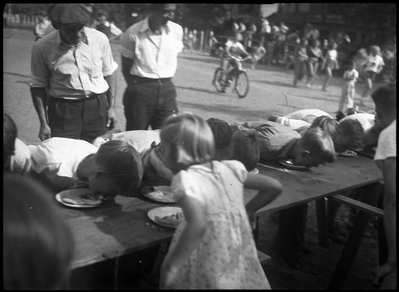 Children competing in a pie eating contest with two adults watching. There are children and other people in the background.