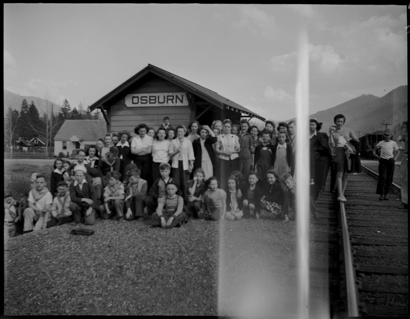 A group of children standing and sitting near a railroad during Boys and Girls Week. A building behind them reads, "Osburn."