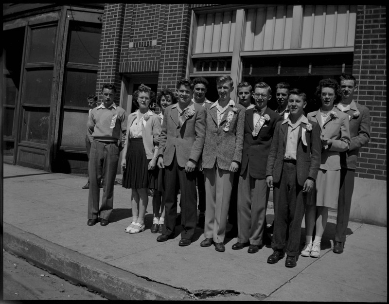 A group of teenagers with ribbons pinned to their shirts during Boys and Girls Week. The teenagers are standing on the sidewalk in front of a brick building.