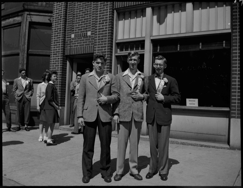Three teenagers with ribbons standing on the sidewalk during Boys and Girls Week. Other teenagers can be seen in the background.