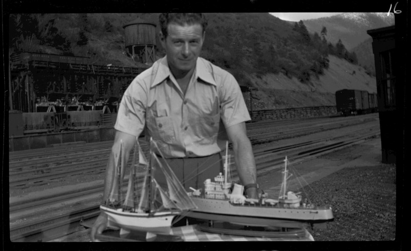 A man stands behind two model ships. There is a railroad in the background.