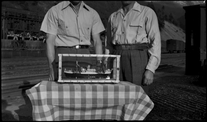 Two men standing behind a model ship. A railroad can be seen in the background.