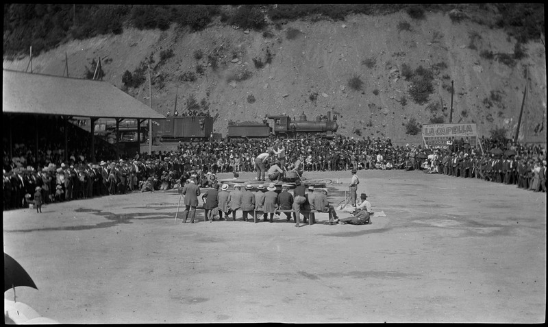 People gathered during a hand-drilling contest at Wallace Park. Several people are seated on a bench in front of the hand-drilling competitors, as the audience crowds around them.