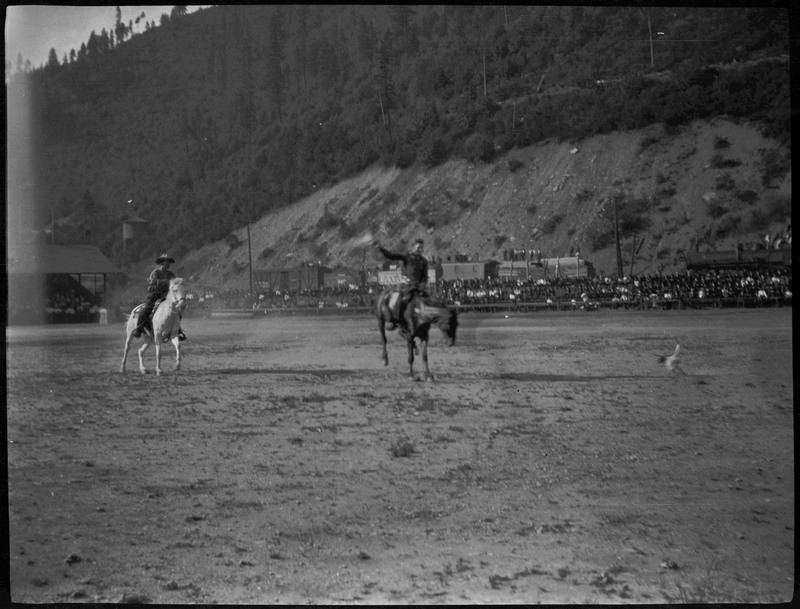 Two people on horseback during a rodeo in Wallace. An audience can be seen in the background.