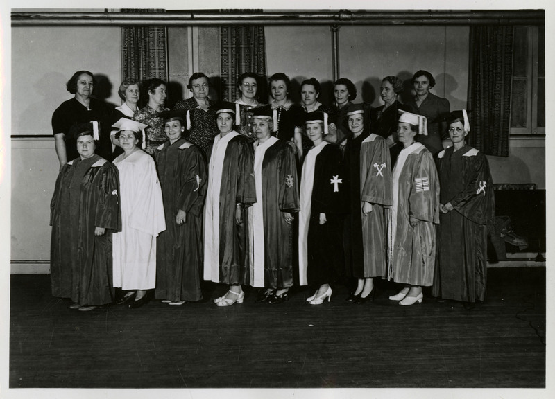 Nine unidentified female graduates wearing caps and gowns stand in front of ten unidentified women.