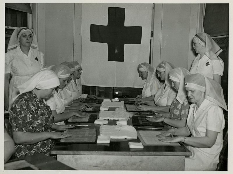 Eight American Red Cross nurses sit at a table with surgical dressing and tools as two other nurses stand watching them. An American Red Cross flag hangs in the background.