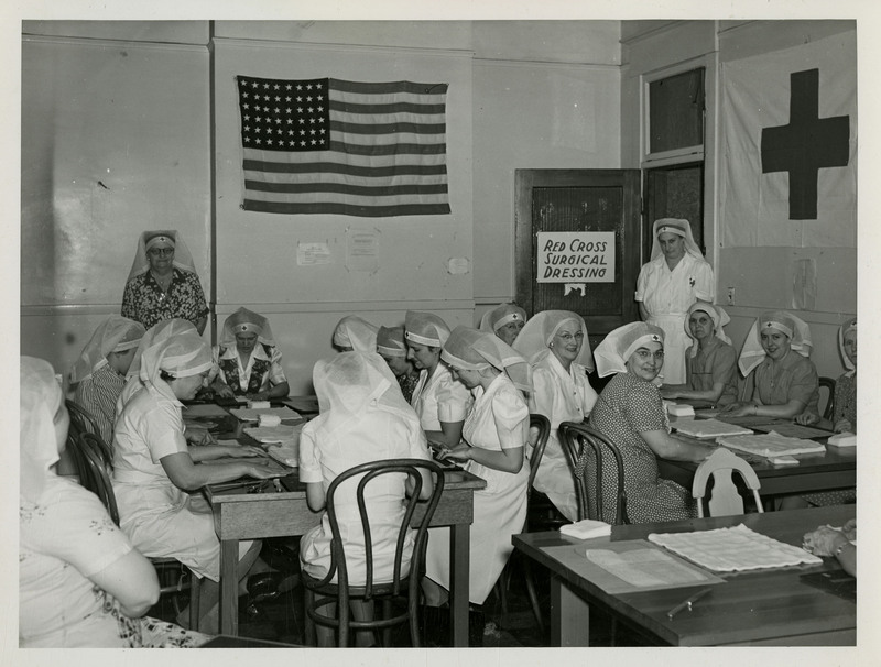 American Red Cross nurses sit at two tables working with surgical dressing and tools as two nurses stand watching them. An American flag and a Red Cross flag hangs on the wall. A sign that reads "Red Cross Surgical Dressing" hangs on the door.
