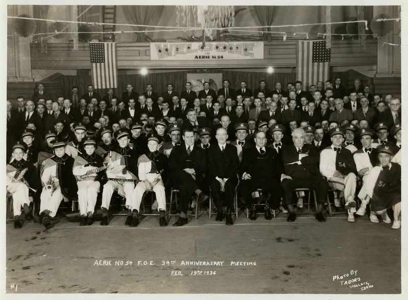 Men and marching band members gather for a photograph in celebration of Aerie #54 Fraternal Order of Eagles' 34th anniversary meeting. A welcome sign hangs between two American flags in the background.