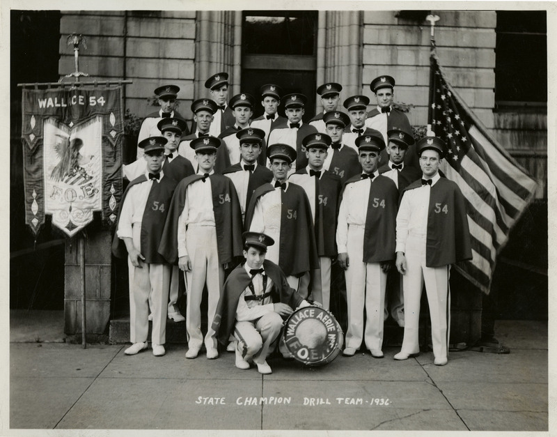Fraternal Order of Eagles Aerie #54 State Champion drill team members pose for a group photo in front of a building. A banner flag with the words "Wallace 54" and "F.O.E." on the right. An American flag on the left.