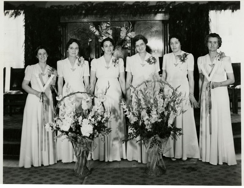 O.E.S. (Order of the Eastern Star of Idaho Grand Chapter) women pose for a photo. Two large flower bouquets placed in front.